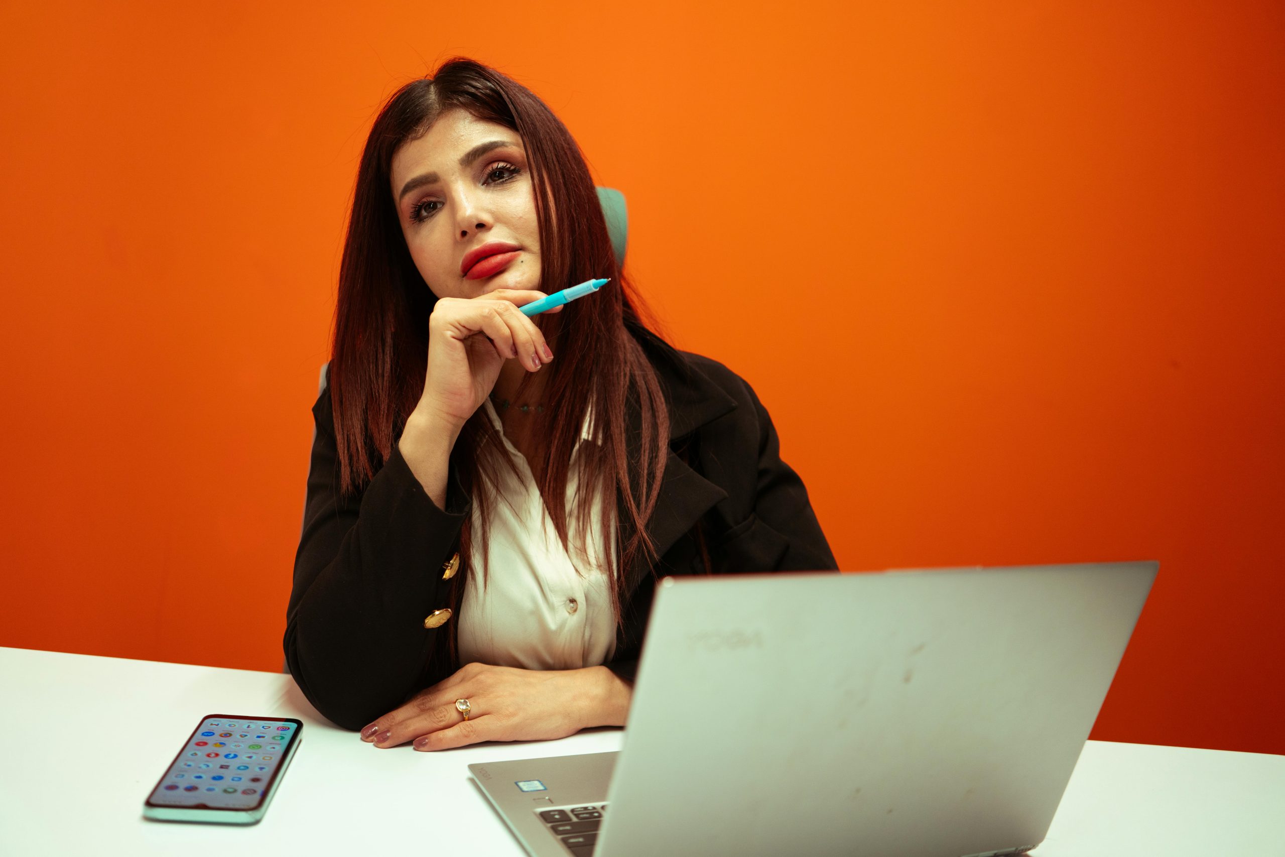 Confident woman in business attire thinking at her desk with laptop and smartphone, against orange background.