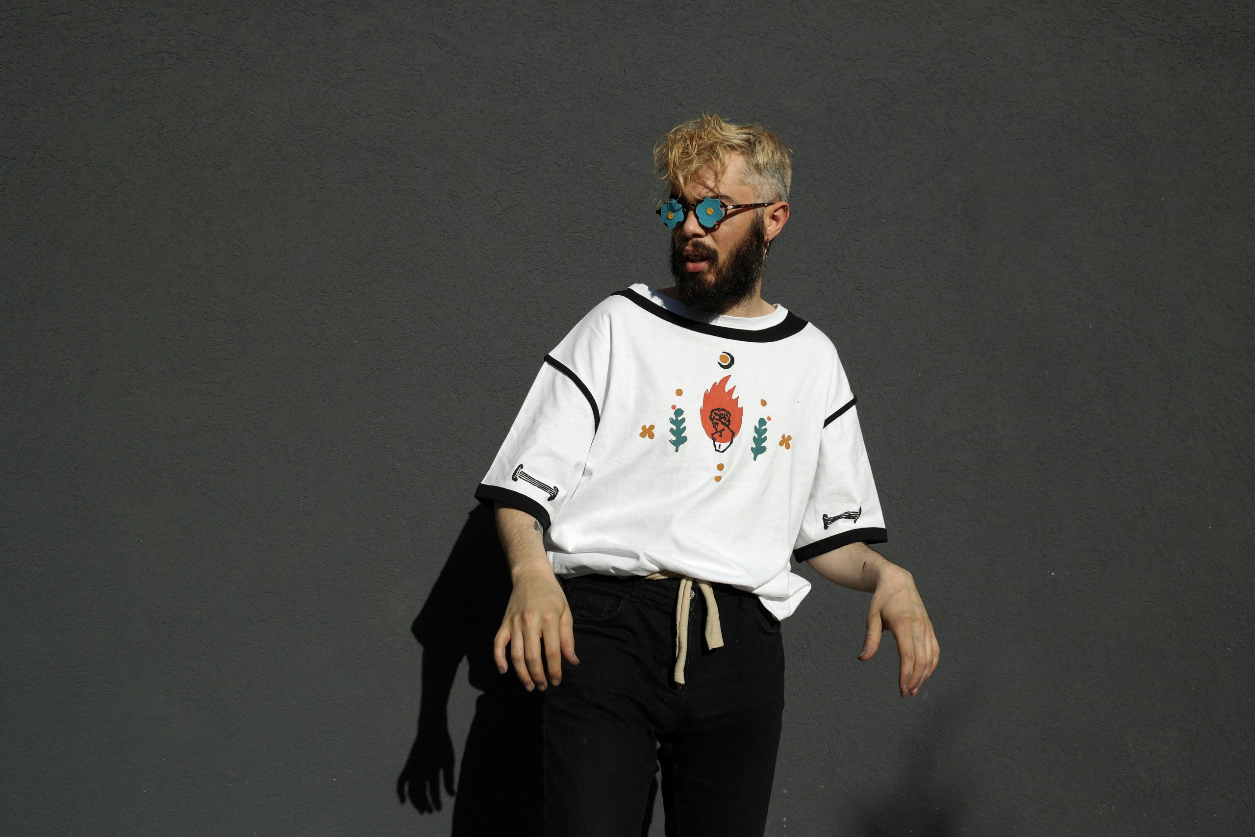 Stylish man with blonde hair and beard wearing distinctive eyeglasses and a graphic tee, casting a shadow on a grey wall.