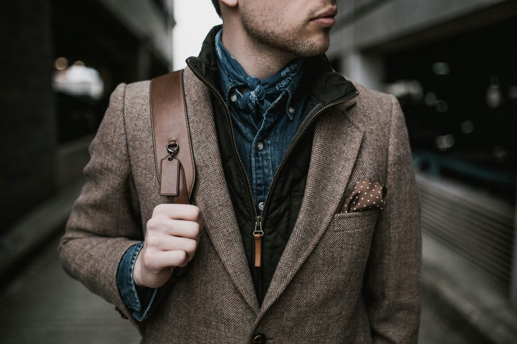 Stylish young man with layered outfit and leather bag posing outdoors in an urban environment.