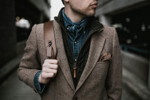 Stylish young man with layered outfit and leather bag posing outdoors in an urban environment.